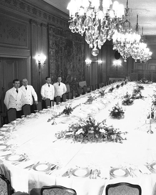Svart-hvitt bilde av servitører i hvite uniformer på siden av et stort selskapsbord med hvite duker og oppdekket til mange gjester